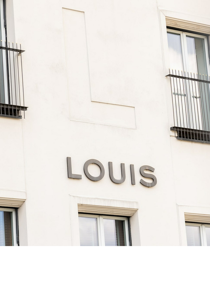 Aussenansicht LOUIS Hotel Muenchen Logo Viktualienmarkt Fassade des LOUIS Hotels in München mit großen Fenstern und Balkongittern.