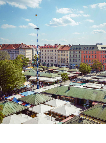 View of the colorful Viktualienmarkt in Munich with maypole, surrounded by historic buildings under a blue sky.
