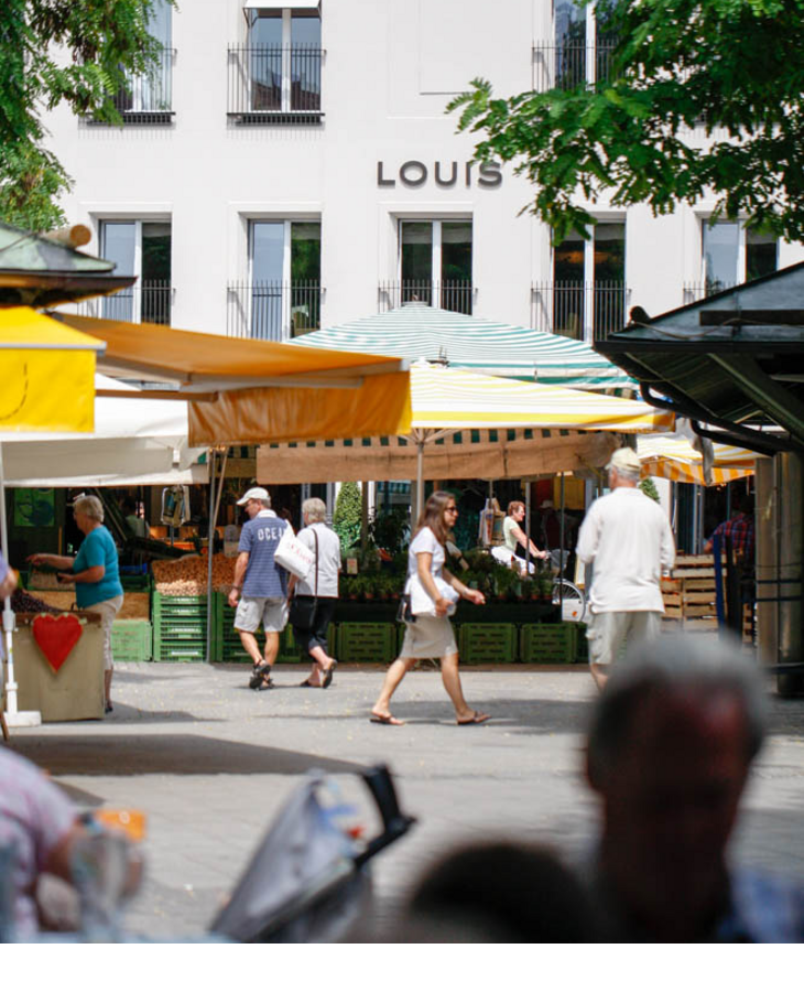Exterior view LOUIS Hotel Munich Viktualienmarkt View of the LOUIS Hotel Munich with colorful market stall in the foreground and passers-by.
