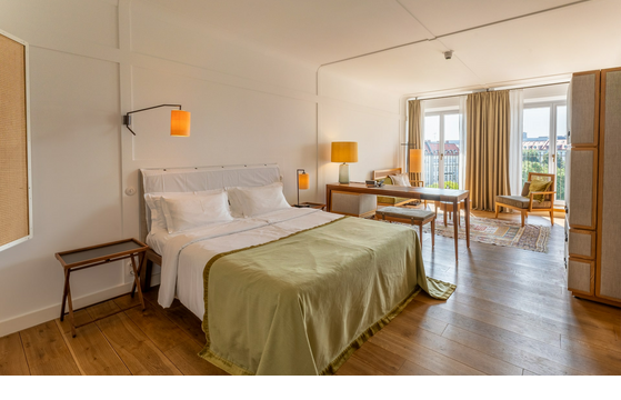 Helles Hotelzimmer mit Doppelbett, Holzboden, Schreibtisch, Sitzbereich und großem Fenster mit Vorhängen.