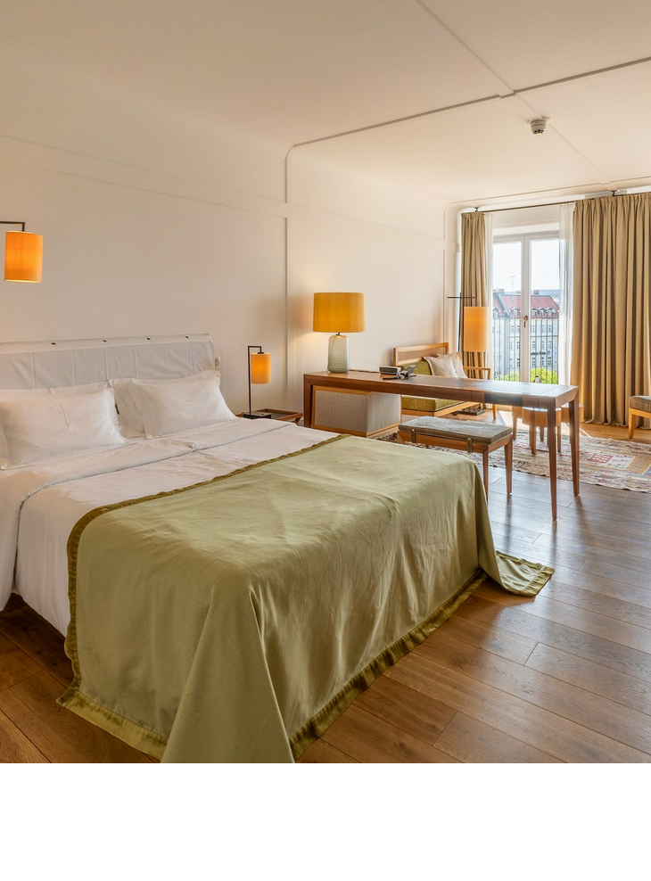 Bright hotel room with double bed, wooden floor, desk, seating area and large window with curtains.