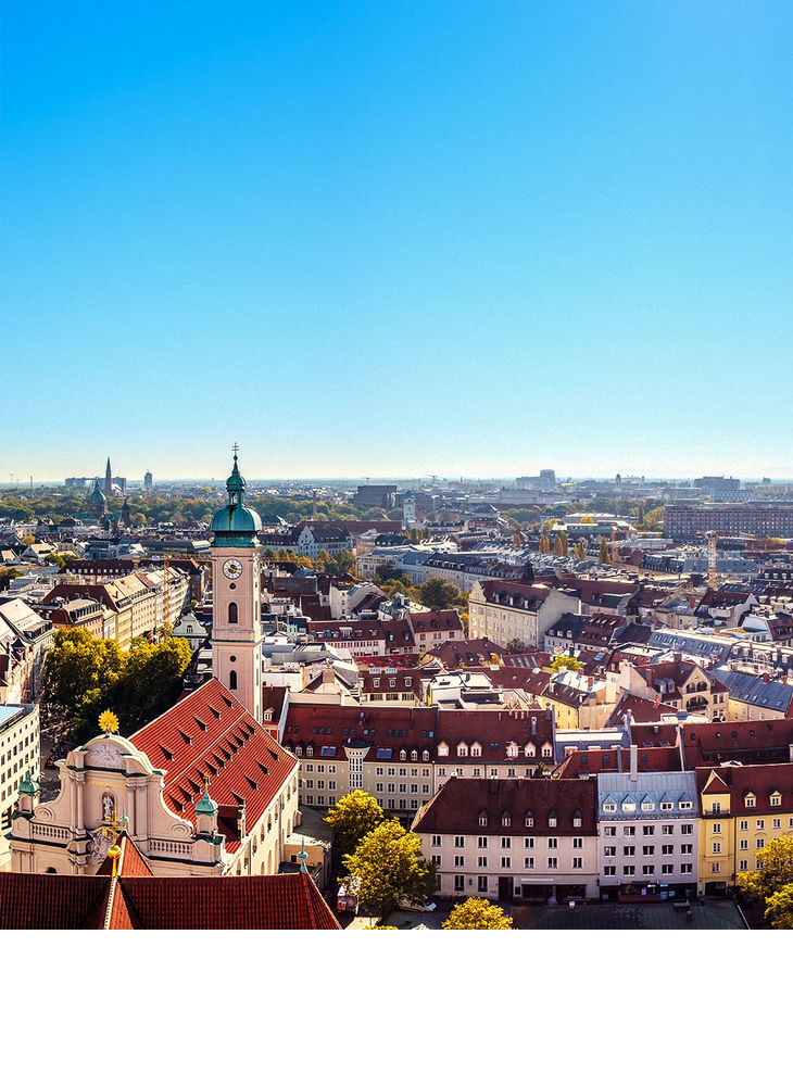 LOUIS Hotel Munich Viktualienmarkt Skyline City view of Munich under a clear sky, with church tower and colorful buildings in the foreground.