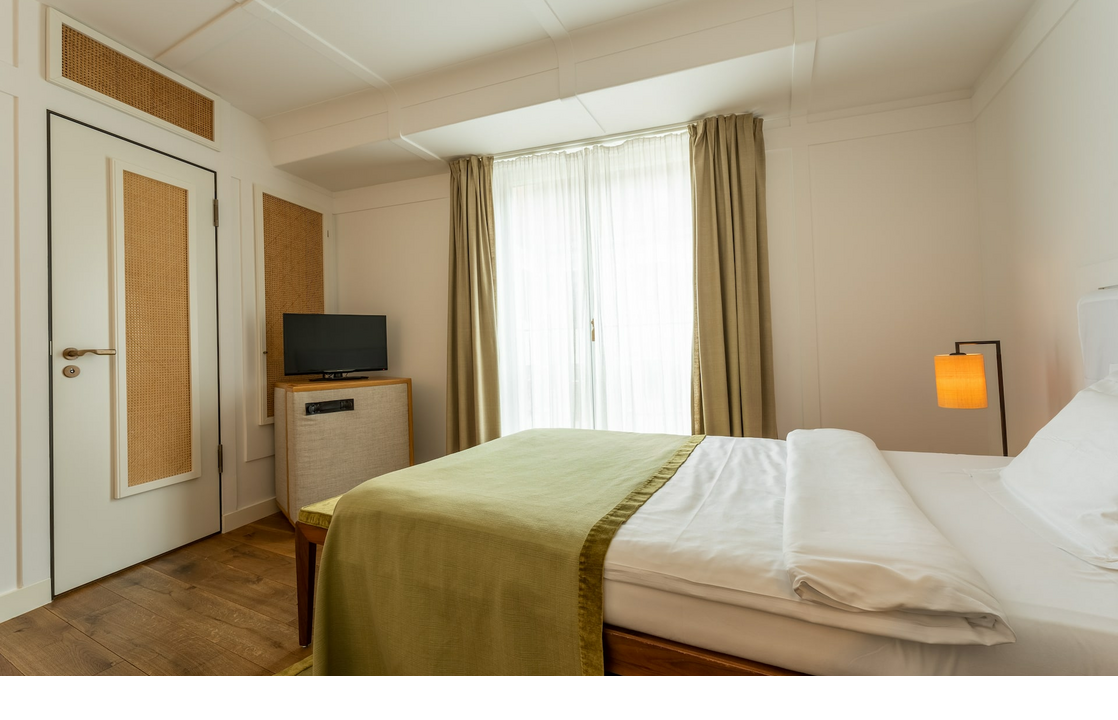 Helles Hotelzimmer mit Doppelbett, grüner Tagesdecke, TV, Lampe und großen Fenstern mit Vorhängen.