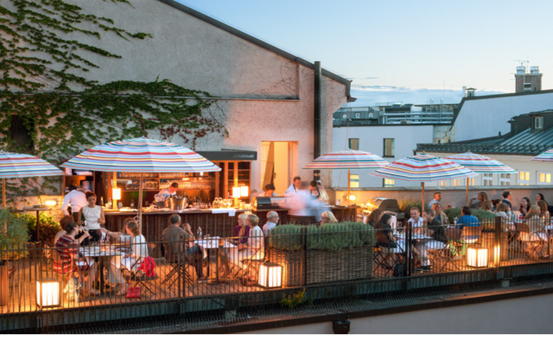 Dachterrasse des LOUIS Hotels in München mit Gästen, bunten Sonnenschirmen und gemütlicher Abendstimmung.