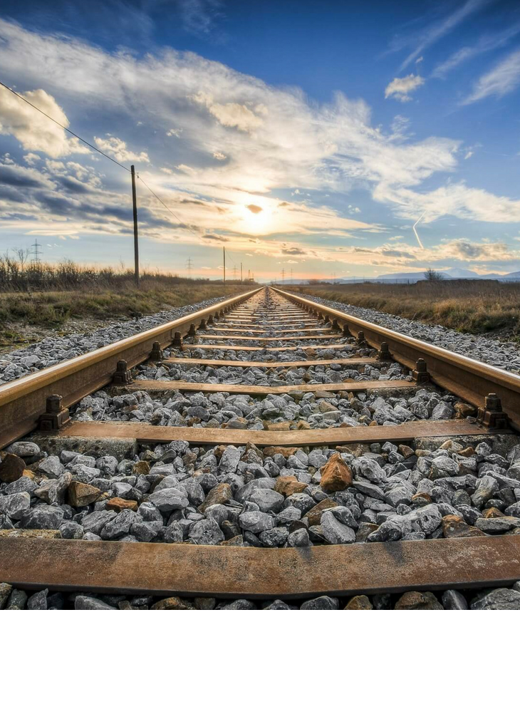 Travelling by German rail to the ATLANTIC Hotels and to the LOUIS Hotel Munich Tracks lead into the sunset, surrounded by gravel and meadows under a partly cloudy sky.