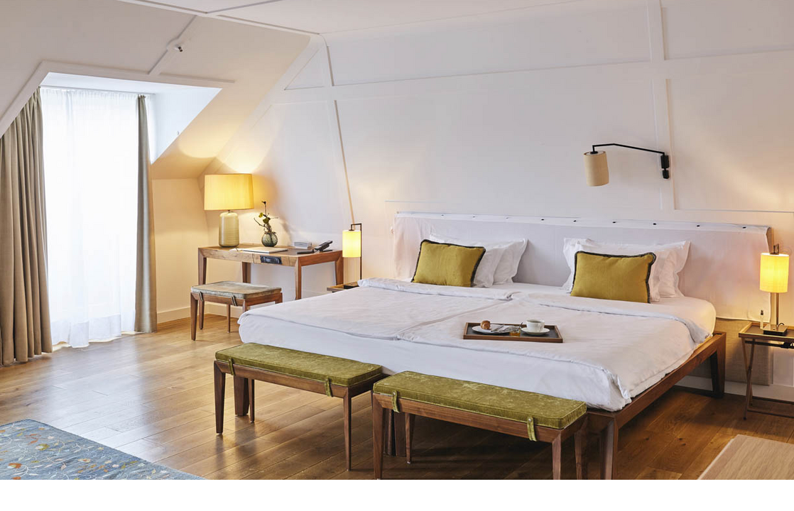 Bright hotel room with large bed, wooden floor, desk, two stools and a window with curtains.