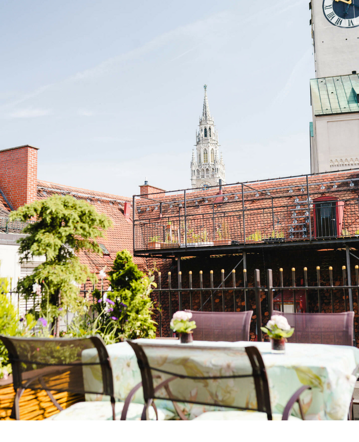 Roof terrace with seating area and plants, view of Munich's church tower under a blue sky.