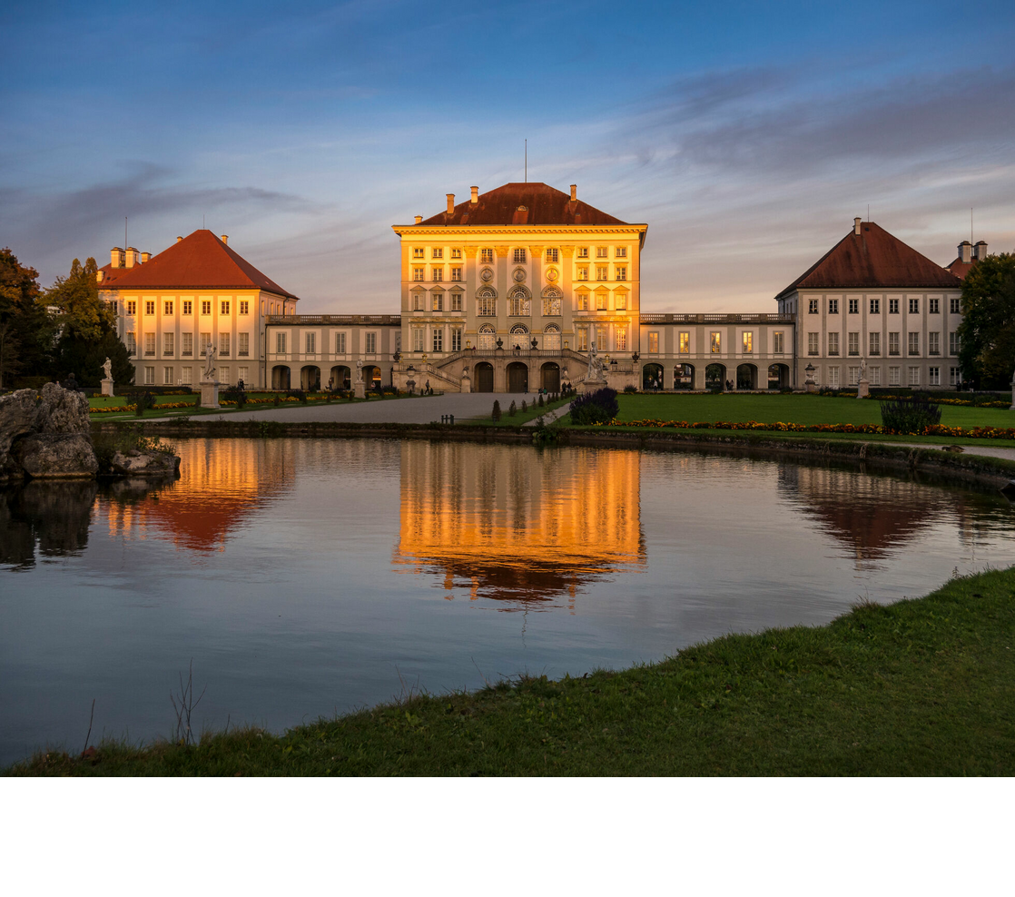 Historisches Schloss mit roten Dächern, umgeben von gepflegtem Garten und Teich, spiegelt sich im Wasser bei Sonnenuntergang.