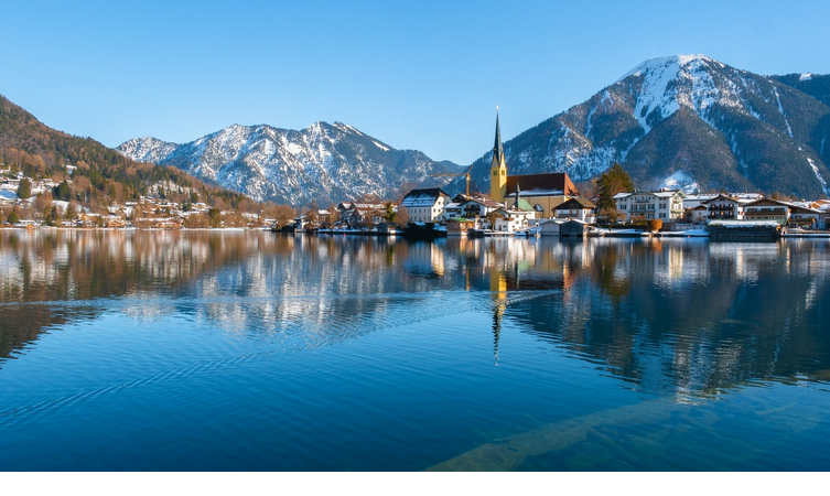 Idyllic lakeside landscape with snow-capped mountains, a village and a church on the shore, reflected in the calm water.