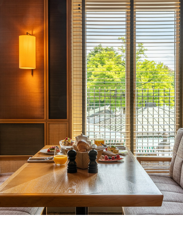 Frühstückstisch im LOUIS Hotel mit frischen Speisen, Saft und Blick auf grüne Bäume durch große Fenster.