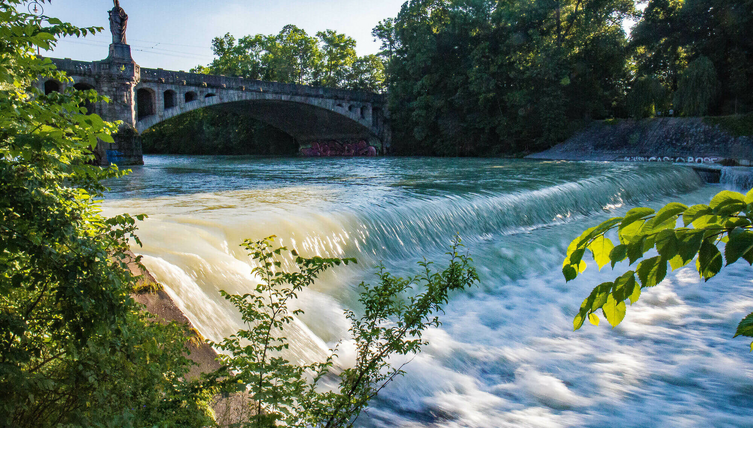 Fluss mit kleinem Wasserfall, darüber eine historische Brücke, umgeben von üppigem Grün und Bäumen.