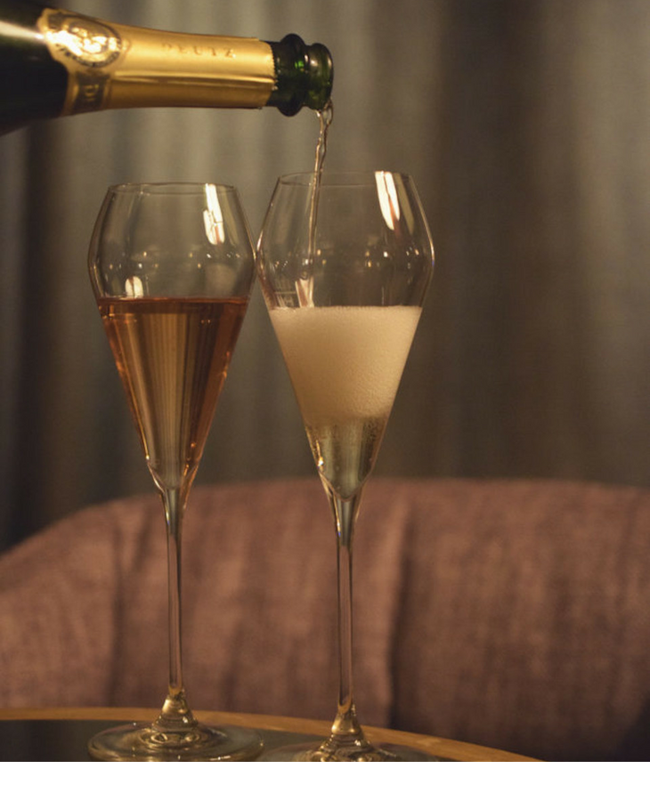 Zwei Sektgläser auf einem Tisch, eines wird mit Champagner gefüllt, im Hintergrund ein gemütlicher Sessel.