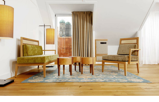 Bright hotel room with stylish wooden furniture, carpet and a view of Munich's Frauenkirche through a large window.