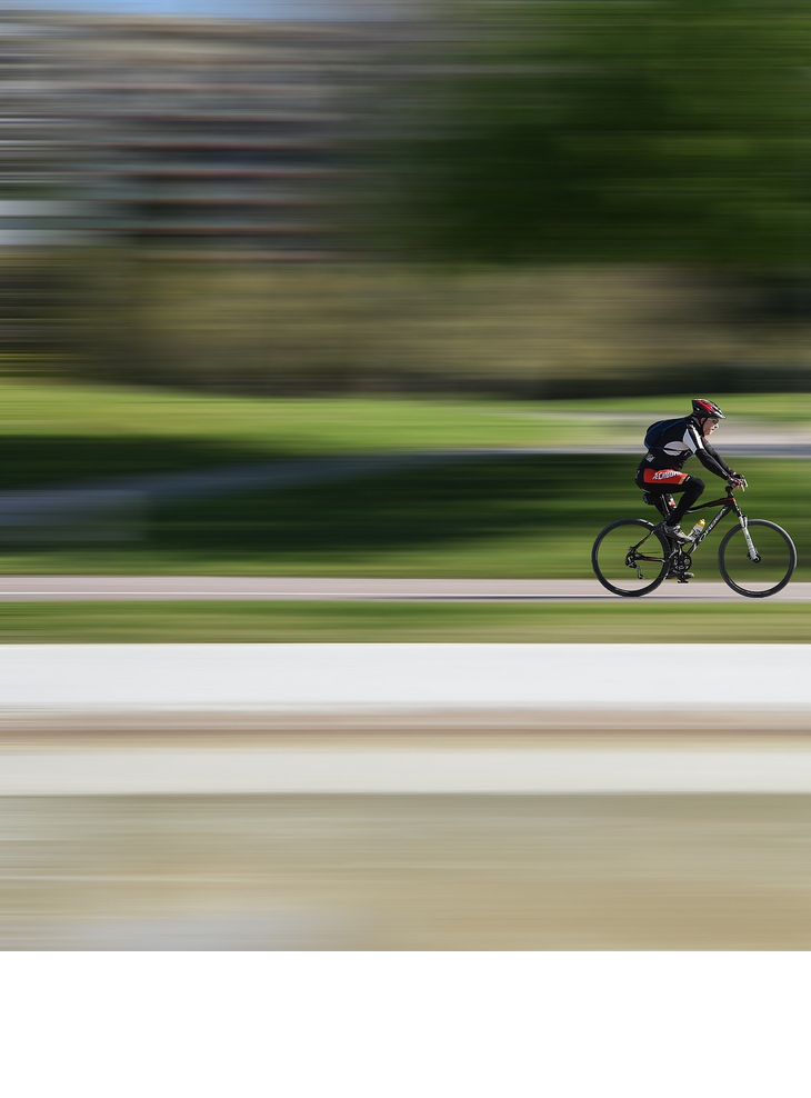 Radfahrer mit rotem Helm fährt schnell auf einem Radweg durch einen grünen Park.