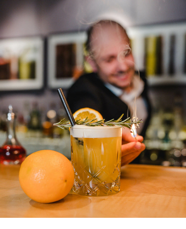 A bartender serves a cocktail with rosemary and a slice of orange, with an orange lying next to it.