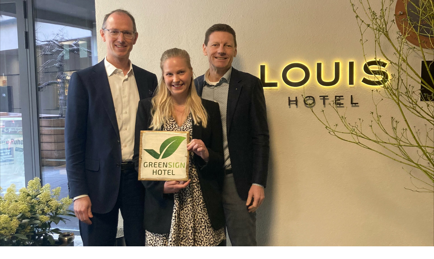 Presentation of the GreenSign certification | The LOUIS Hotel Munich Three people in front of the LOUIS Hotel logo hold a Greensign certificate, which recognizes sustainability.
